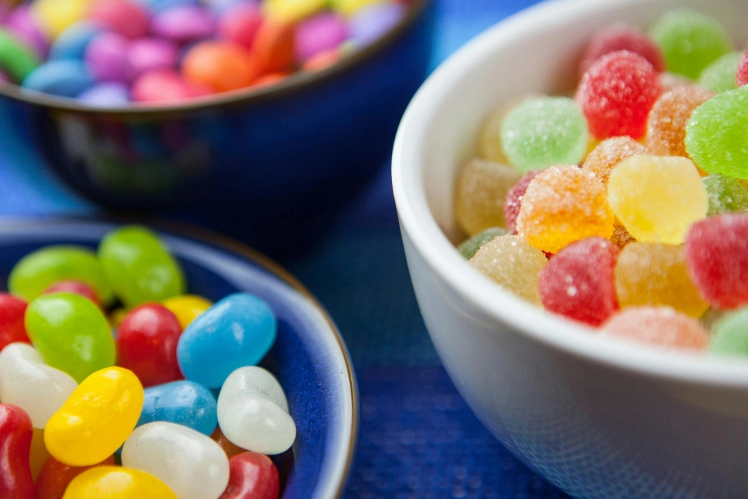Colorful candies and gummy sweets arranged in white bowls on blue surface.