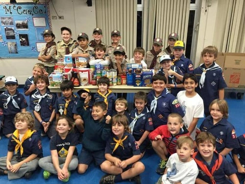 Large group of children in scout uniforms seated together in a gymnasium or indoor facility.