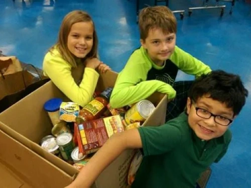 Three children in bright colored shirts posing with donation box in blue gymnasium setting.