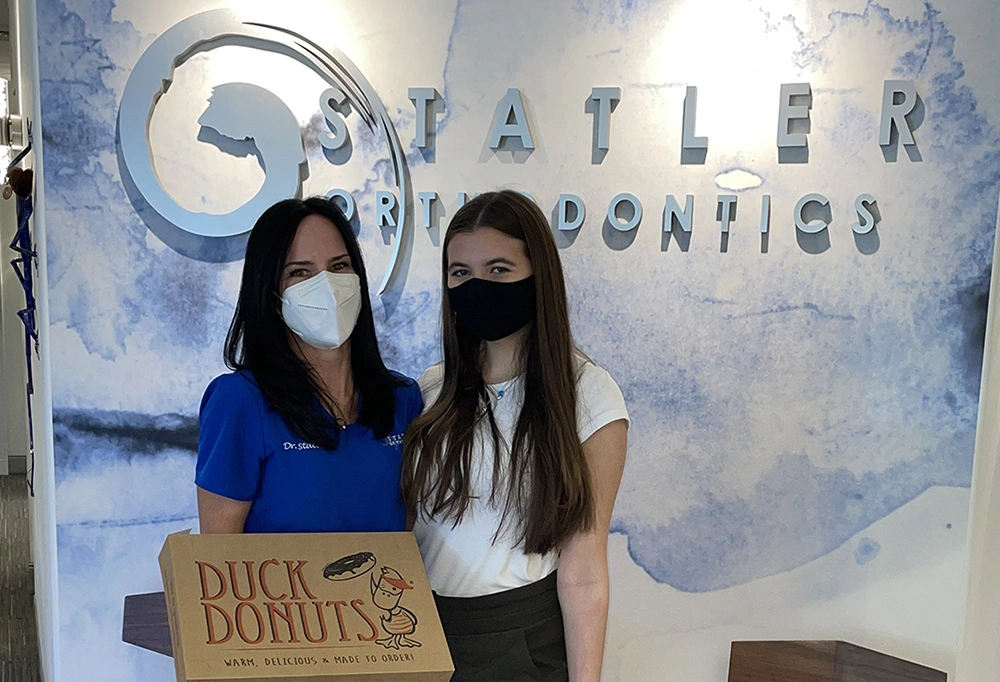 Two women wearing masks hold Duck Donuts box in front of Statler Orthodontics signage.