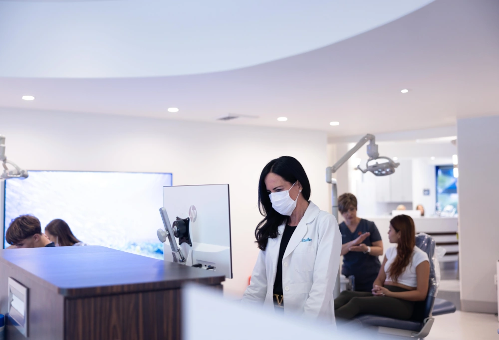 Orthodontist in mask and white coat stands in modern dental office with patients seated in waiting area.