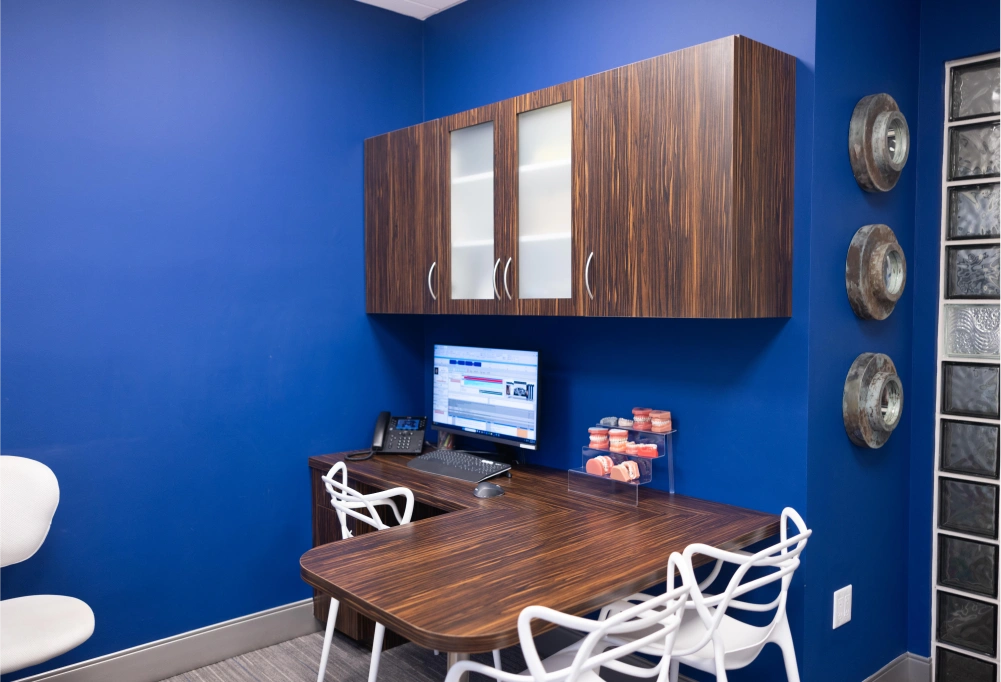 Modern orthodontics office with blue walls, wooden desk, computer setup, and professional cabinetry.