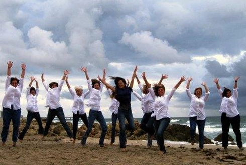 Group of people in white shirts jumping with arms raised on beach under cloudy sky