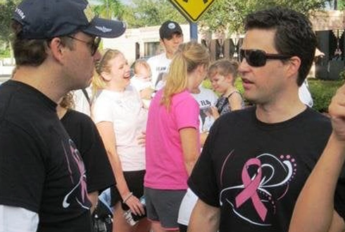 Two men wearing pink ribbon shirts converse at an outdoor community event with children nearby.