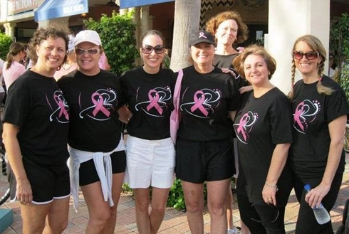 Group of seven people wearing matching black t-shirts with pink logo at outdoor event.