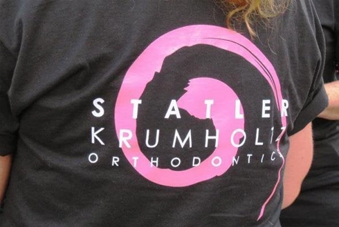 Person wearing black Statler Kumholia Orthodontics t-shirt with pink circular logo design.