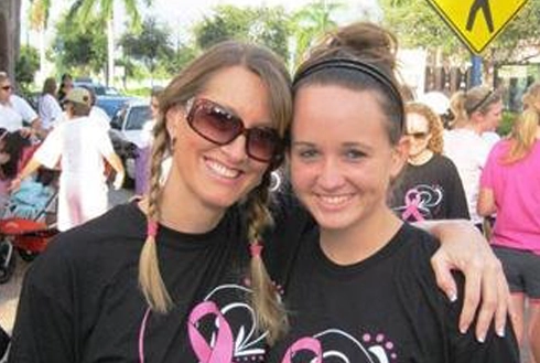 Two smiling women at outdoor event wearing black shirts with pink graphics, sunny day with crowd in background.