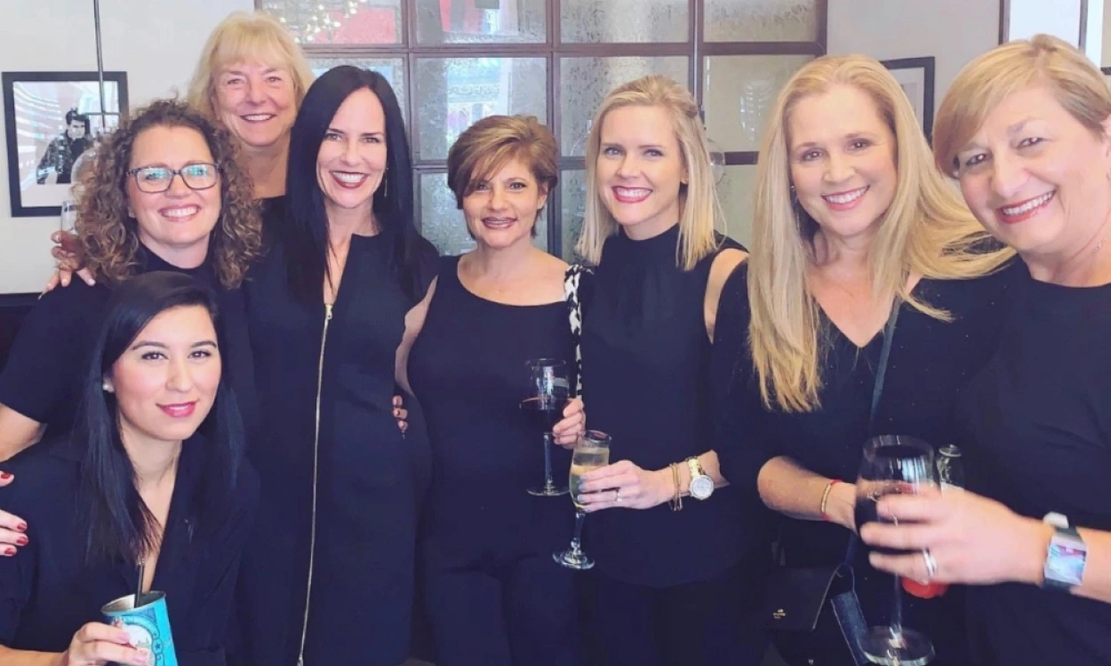 Group of women in dark clothing at an indoor event, smiling and holding drinks together.