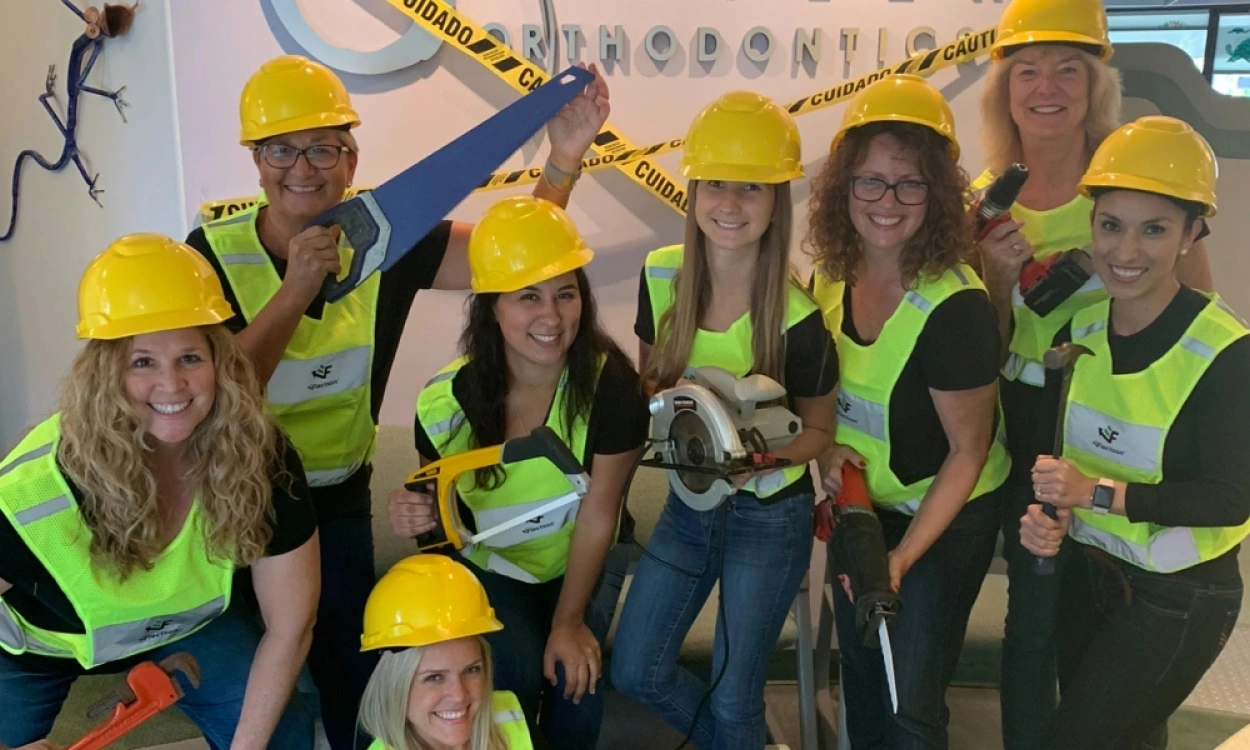 Group of people wearing yellow hard hats and green safety vests posing together indoors.