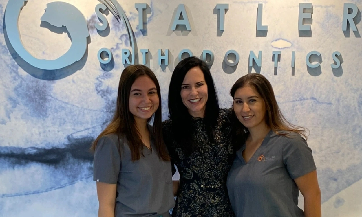 Three women smile in front of Statler Orthodontics office signage on wall.