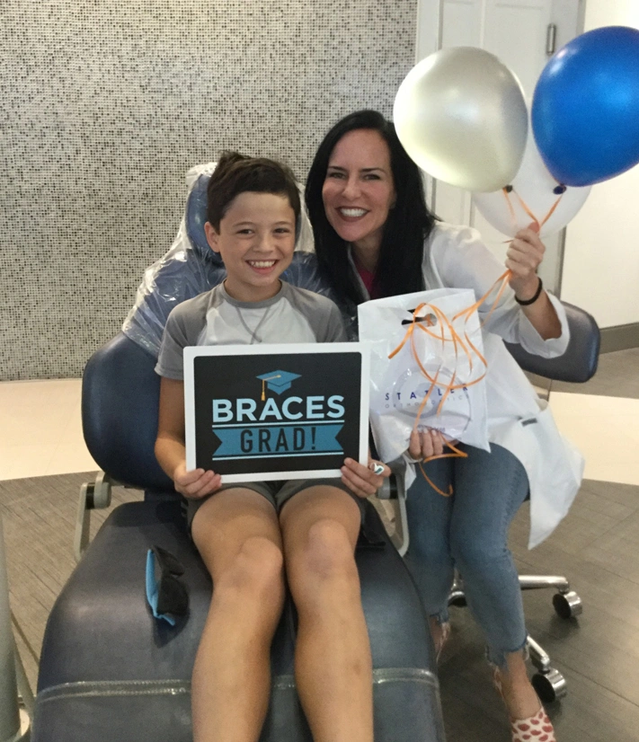 Young boy and woman holding braces sign and certificate with balloons in orthodontic office.