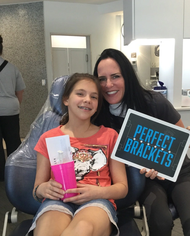 Woman and girl smile together in orthodontic office, holding book and tablet displaying "Perfect Brackets."