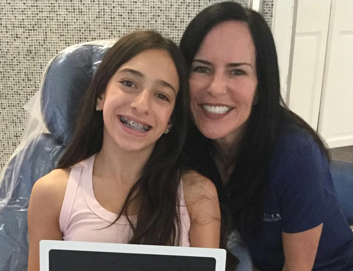 Two smiling women posing together indoors with a laptop, orthodontic office setting.