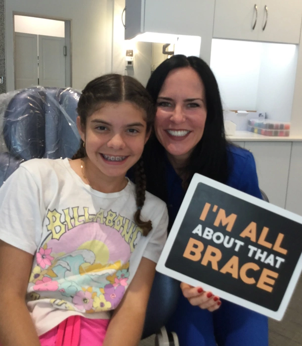 Two people smiling at camera, one holding "I'm All About Grace" sign in orthodontic office.