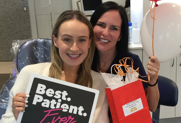 Two women smiling at orthodontic office holding "Best Patient" sign and gift bag.