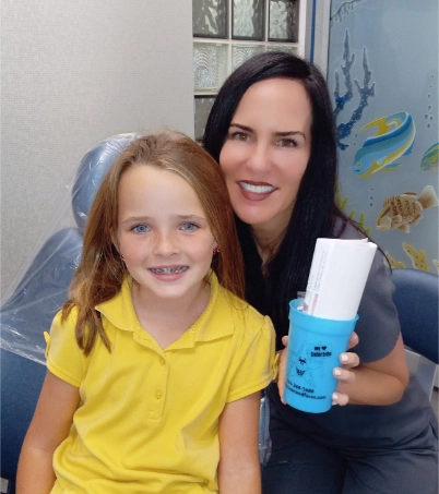 Woman and young girl smiling at camera, girl wearing yellow shirt, woman holding blue product bottle in orthodontic office.