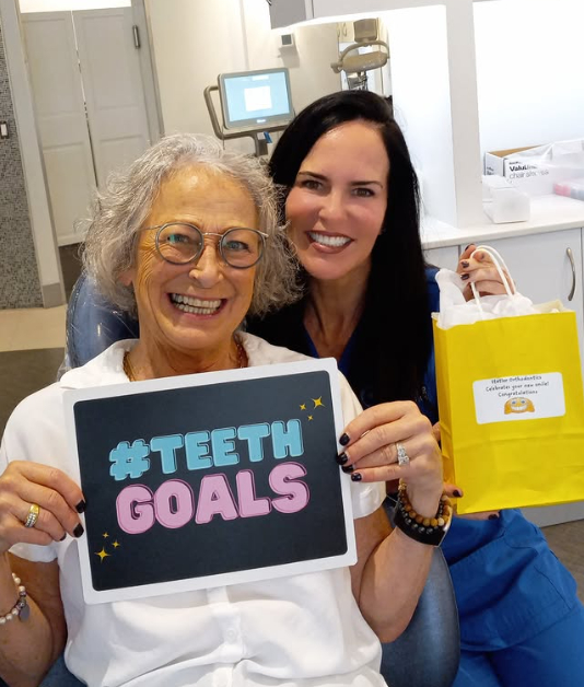 Two women smiling at camera holding tablet displaying "#TEETH GOALS" in orthodontic office setting.