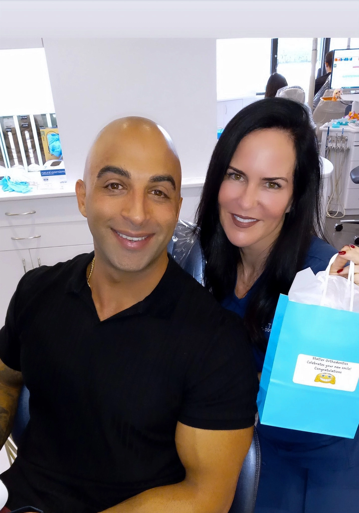 Two smiling professionals pose together in a bright orthodontic office setting with shelving visible behind them.