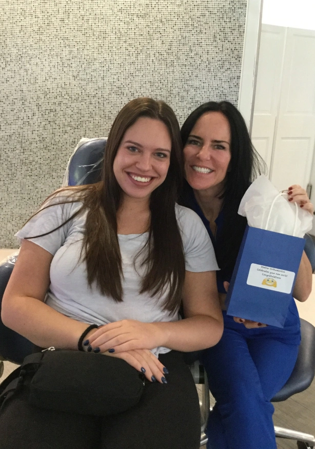 Two women smiling at camera in dental office setting with blue folder visible.