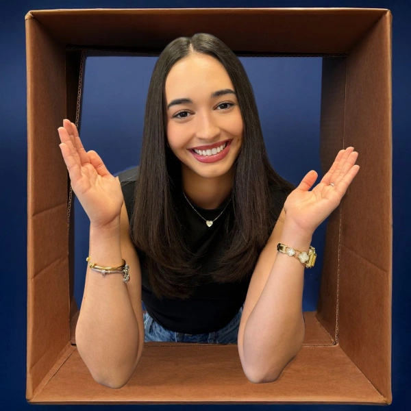 A smiling young woman with long, dark hair poses playfully inside a large, open cardboard box frame against a blue background, raising both hands with open palms. Below the image, text reads "Naomi" and "Scheduling Coordinator".